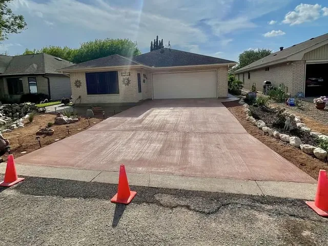 A poured concrete driveway in a red-brown color with a broom finish, running down to the street with traffic cones set out at the curb
